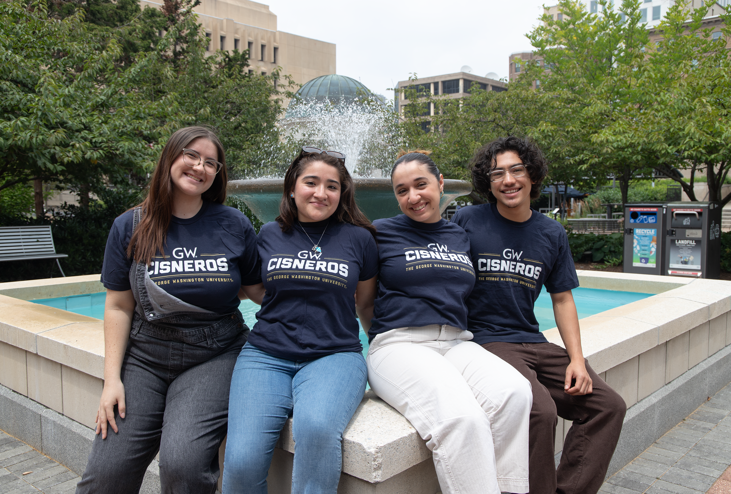 Four Cisneros Scholars sit by a fountain facing the camera and wearing navy shirts