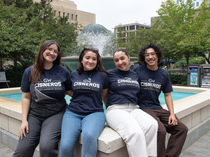 Four Cisneros Scholars sit by a fountain facing the camera and wearing navy shirts